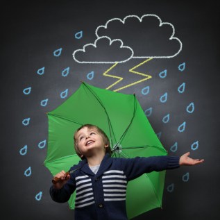 Young happy child singing and dancing holding an umbrella standing in front of a chalk drawing of a rain and lightning storm on a school blackboard